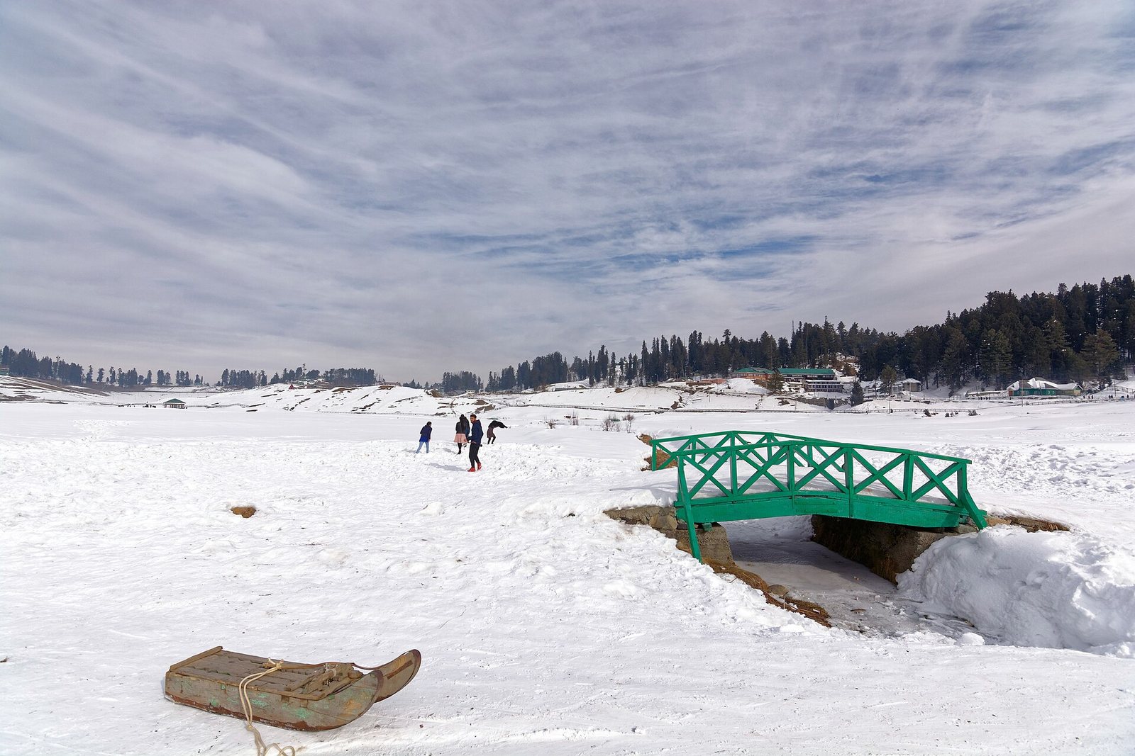 Snowfields at Gulmarg with a small green footbridge and pine forest beyond